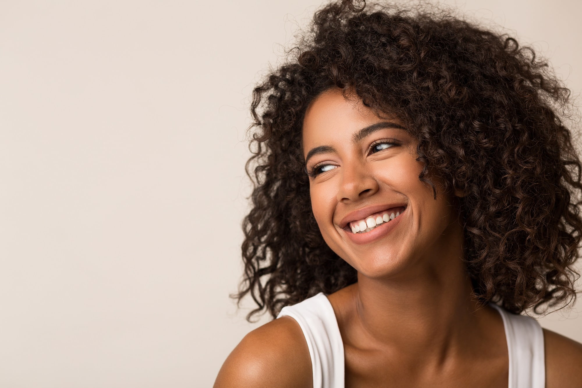 A woman with curly hair smiles brightly, looking off to the side, against a light beige background - professional teeth whitening A woman with curly hair smiles brightly, looking off to the side, against a light beige background - professional teeth whitening