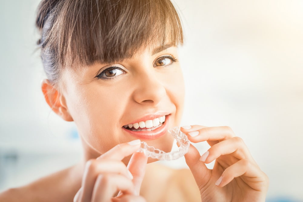 A woman smiles while holding a clear dental aligner near her mouth, ready to use it - Invisalign treatment A woman smiles while holding a clear dental aligner near her mouth, ready to use it - Invisalign treatment