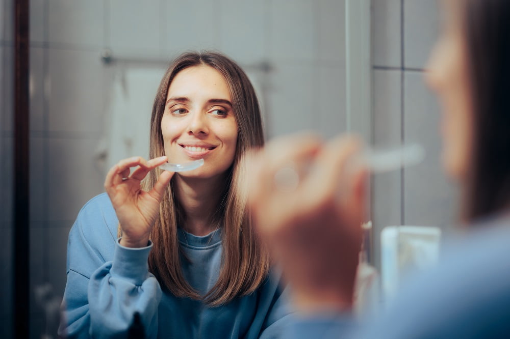 A woman smiles as she brushes her teeth in front of a bathroom mirror - dentures A woman smiles as she brushes her teeth in front of a bathroom mirror - dentures