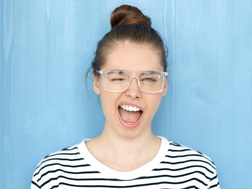 A woman wearing glasses and a striped shirt smiles with a playful, open-mouthed expression against a blue background - veneers and dental laminates A woman wearing glasses and a striped shirt smiles with a playful, open-mouthed expression against a blue background - veneers and dental laminates