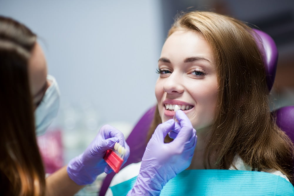 A dental professional holds a color-matching shade guide next to a smiling patient's teeth to determine the ideal shade for treatment - dental crowns and bridges A dental professional holds a color-matching shade guide next to a smiling patient's teeth to determine the ideal shade for treatment - dental crowns and bridges