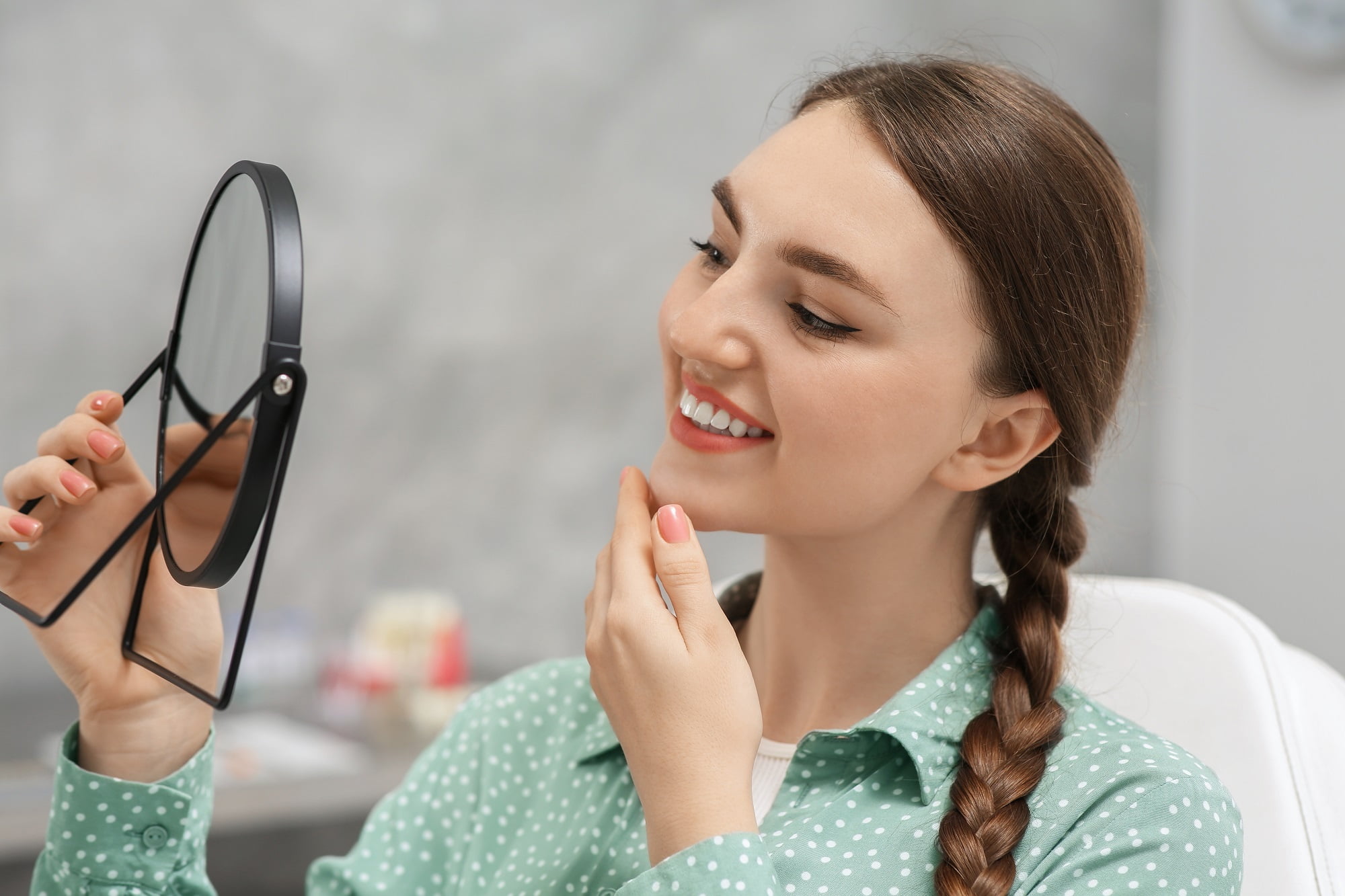 Young woman smiling while examining her teeth in a handheld mirror after a dental checkup - dental implants Young woman smiling while examining her teeth in a handheld mirror after a dental checkup - dental implants