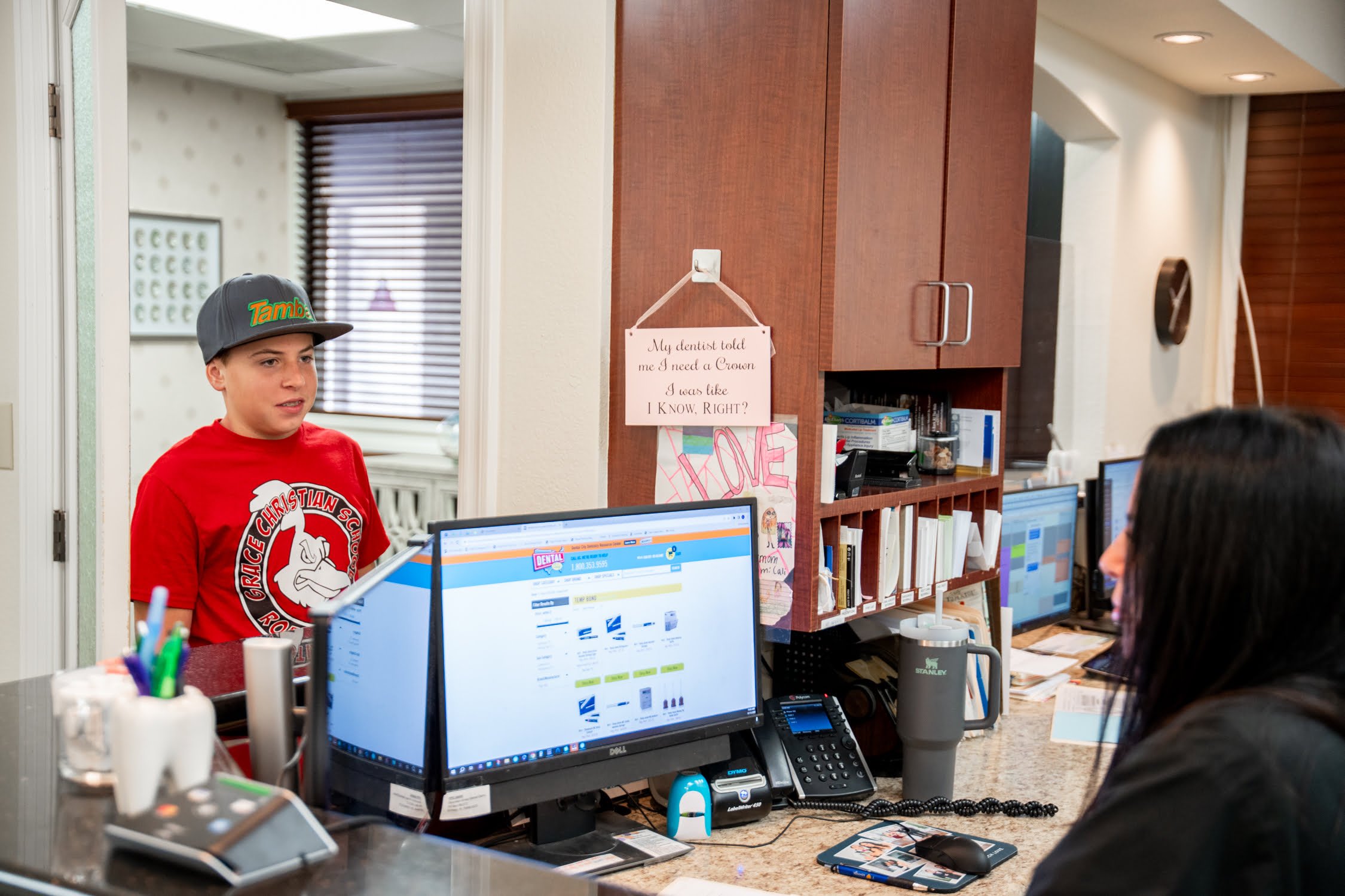 A young boy in a red shirt and cap stands at the reception desk, speaking with a receptionist in a dental office - smile makeover A young boy in a red shirt and cap stands at the reception desk, speaking with a receptionist in a dental office - smile makeover