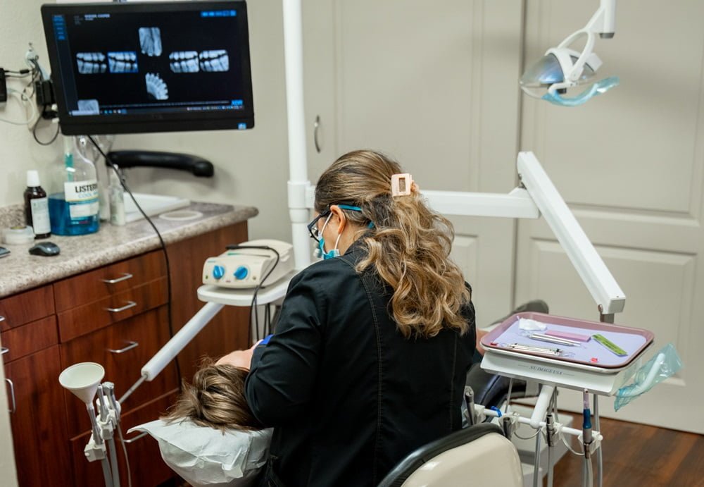 A dental professional performs a procedure on a patient in a dental chair while an x-ray image of teeth is displayed on a screen in the background - suresmile clear aligners A dental professional performs a procedure on a patient in a dental chair while an x-ray image of teeth is displayed on a screen in the background - suresmile clear aligners