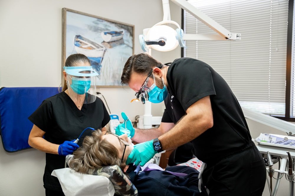 Assistant perform a procedure on a patient in a dental office, both wearing masks and protective eyewear - dental crowns and bridges Assistant perform a procedure on a patient in a dental office, both wearing masks and protective eyewear - dental crowns and bridges