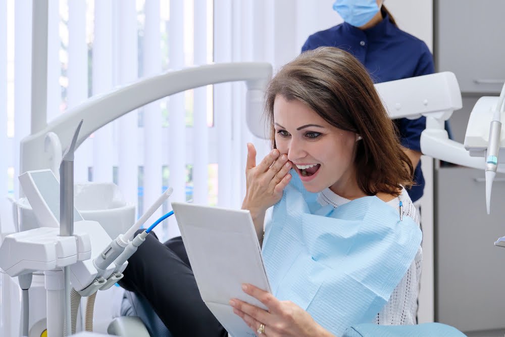 A woman in a dental chair smiles as she looks at her reflection in a hand mirror, pleased with her dental results - Cosmetic dentistry A woman in a dental chair smiles as she looks at her reflection in a hand mirror, pleased with her dental results - Cosmetic dentistry