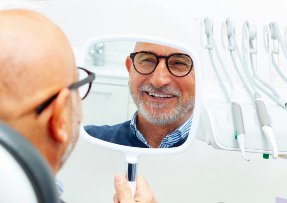 Senior Man Admiring His Smile in Mirror – Dentist in Long Beach CA An older man smiling at his reflection in a handheld mirror at a dental clinic, appreciating his rejuvenated and healthy smile – Dentist in Long Beach CA.