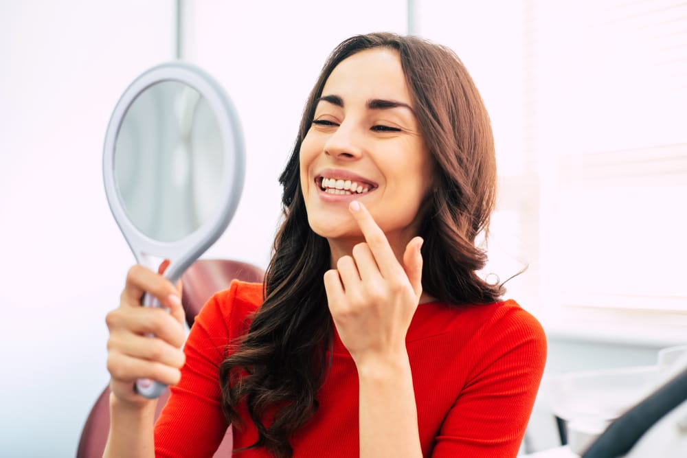 Woman Admiring Her Smile in Mirror – Cosmetic Dentistry in Long Beach CA A woman in a red top smiles as she looks in a handheld mirror at the dental office, appreciating her enhanced and radiant new smile – Cosmetic Dentistry in Long Beach CA.