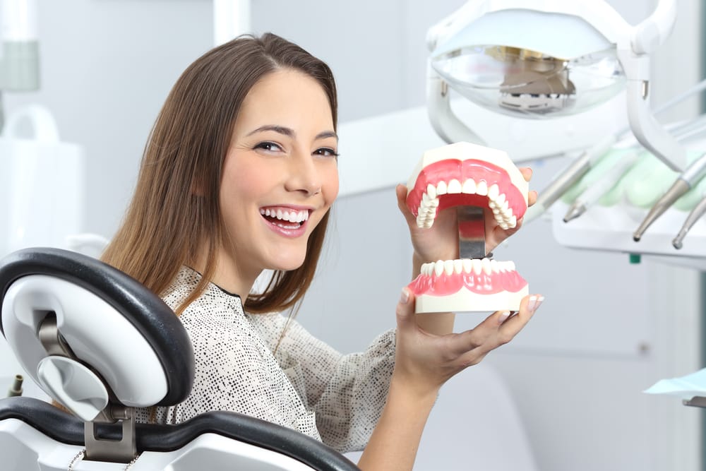 Smiling Woman Holding Denture Model – Long Beach Dentures A cheerful woman sits in a dental chair and holds a full denture model, representing quality dentures and personalized dental care – Long Beach Dentures.