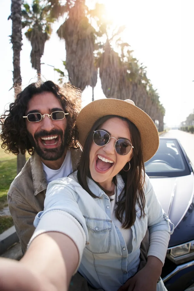 A cheerful couple enjoys a sunny day outdoors, smiling widely as they take a selfie near palm trees and a parked car. Their joyful expressions and casual outfits capture a carefree California vibe, perfect for lifestyle or dental care promotions focused on bright, confident smiles.